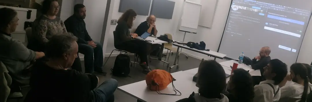 Una docena de personas sentadas formando un círculo en una sala de Las Naves de València, aparentemente debatiendo sobre Twitter, BlueSky y Mastodon, por lo que aparece en una proyección sobre la pared en el fondo de la sala.