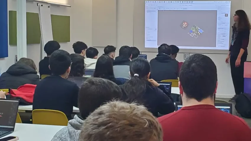 Un grupo de personas con ordenadores portátiles en un aula prestan atención a una persona explicando el motor de Godot sobre una pantalla proyectada en la pared.