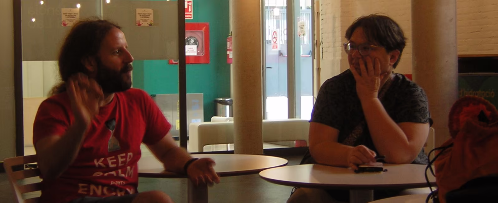 Dos personas que parecen estar participando en un debate más grande. La de la izquierda parece estar hablando en ese momento, realizando gestos con las manos. La persona a la derecha muestra un gesto de atención a lo que se está diciendo.