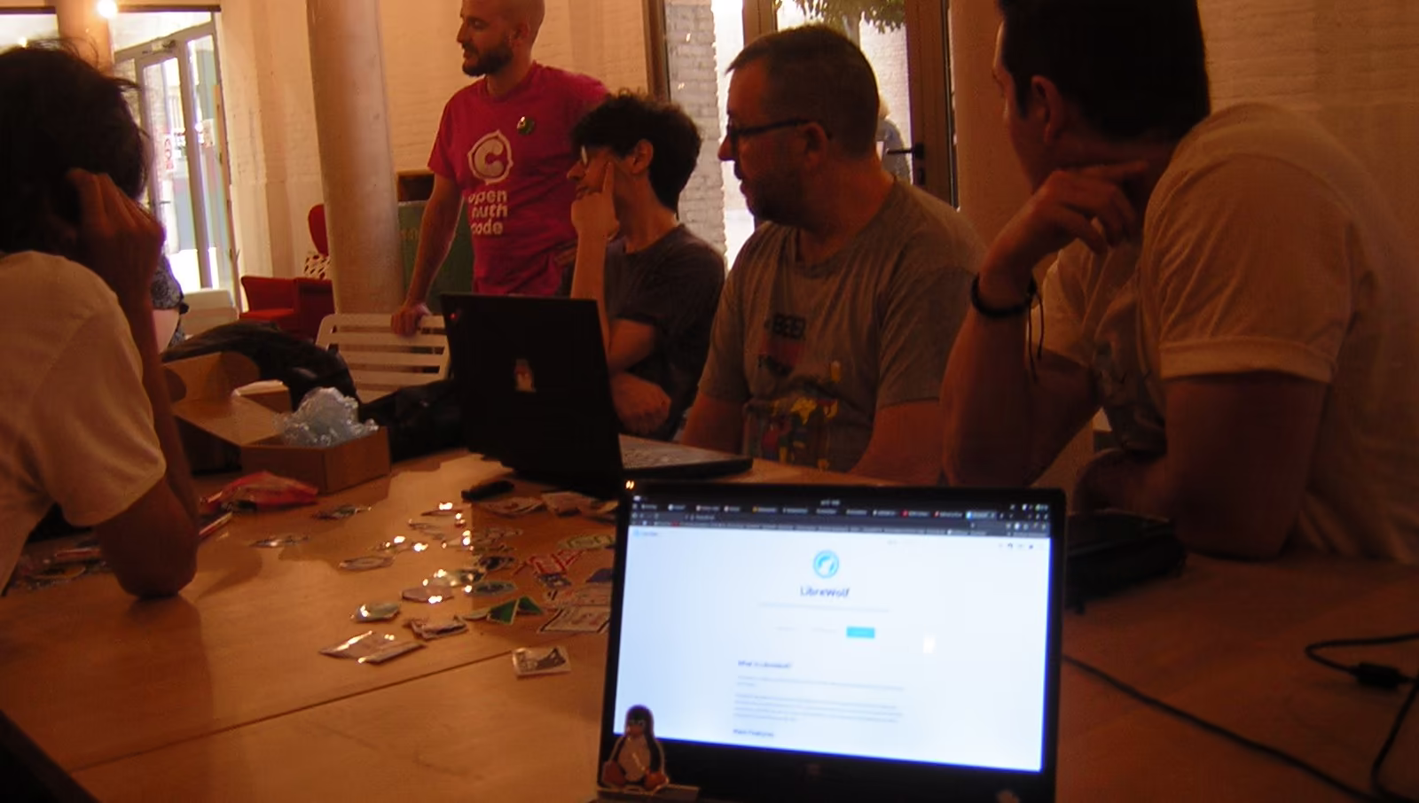 Múltiples personas que parecen estar escuchando a alguien comentar algo. Sobre la mesa, se ven pegatinas de software libre. En primer plano, hay un portátil con una pestaña del navegador abierta mostrando el proyecto LibreWolf.